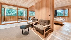 Ein modernes Zimmer mit Holzwänden und -böden, großen Fenstern mit Blick auf einen Wald, einer Couch, einem kleinen Tisch und einem Bett.