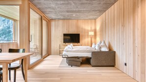 A modern bedroom with a wooden floor, a bed with white bedding, a TV mounted on the wall, and a window providing a view of trees. - Studa 29 - Studa 1 in Brand (Brand)