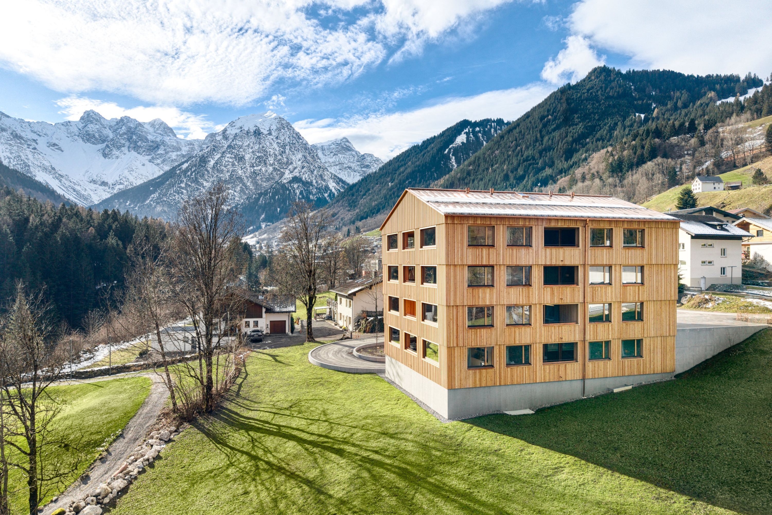 A modern building with a wooden facade, surrounded by a well-maintained lawn and a mountain backdrop.