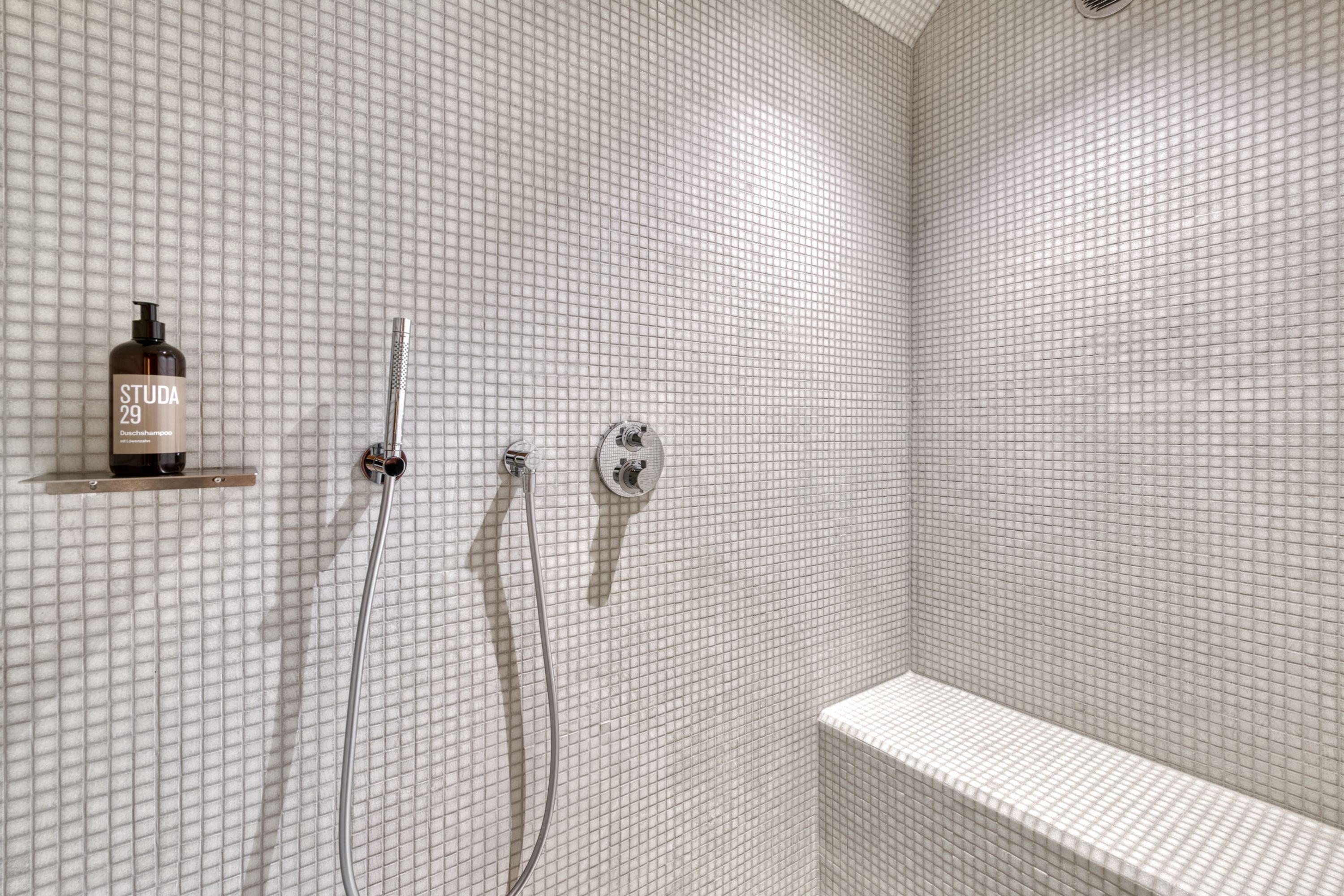 A shower area with a tiled wall, a showerhead, a handheld shower, and a bottle of shampoo on a shelf.