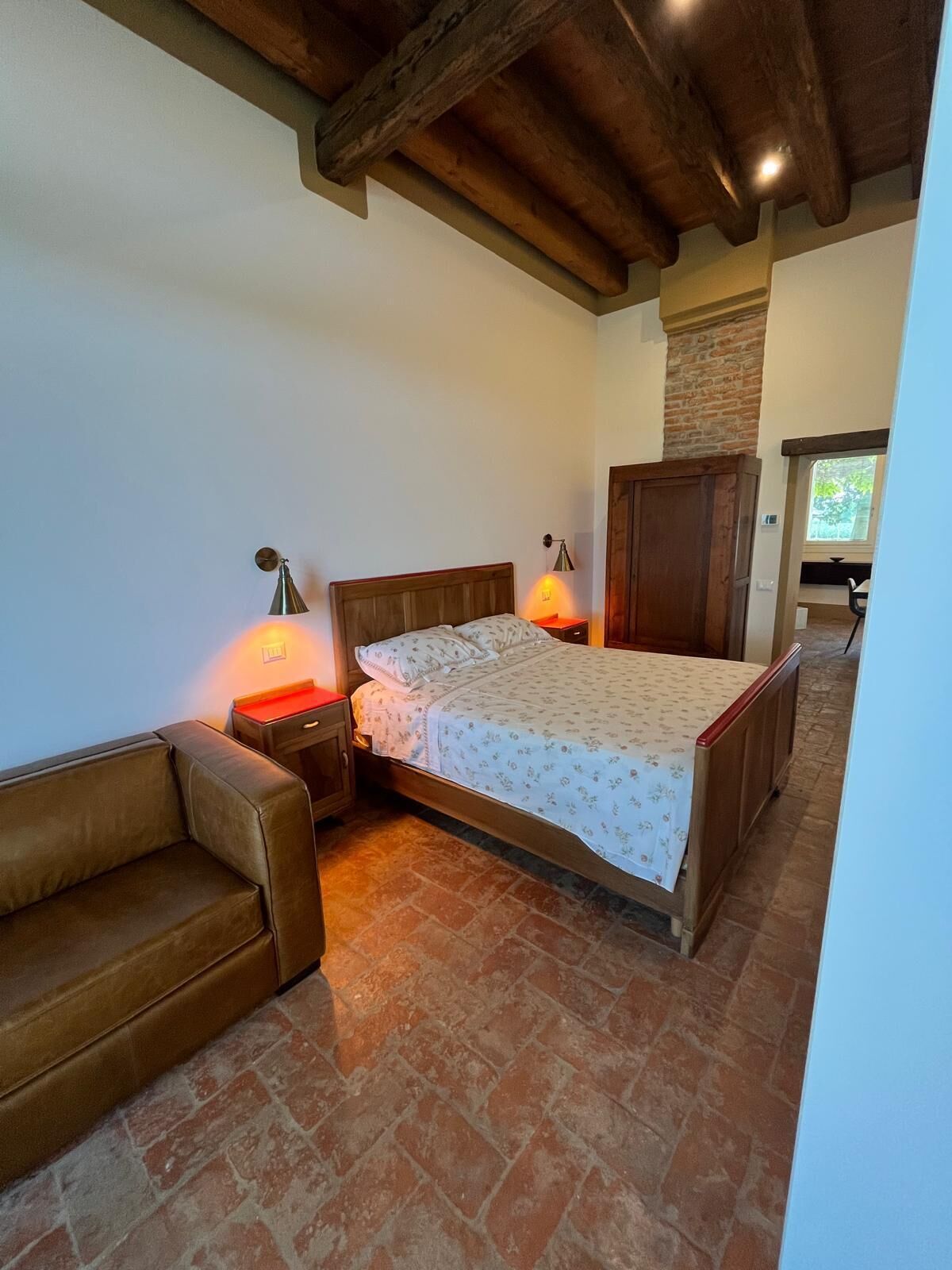 A bedroom with a wooden bed, a nightstand with a lamp, a leather sofa, and a brick floor.