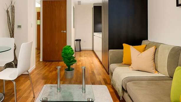 A modern living room with a glass coffee table, a sofa with cushions, a dining area with a glass table and chairs, and a wooden door leading to another room. - Still Life Moorgate Deluxe (London)