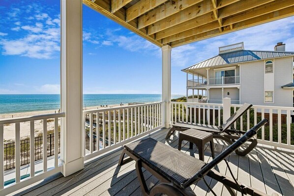 A wooden deck with lounge chairs overlooking a beach and a house with a balcony.