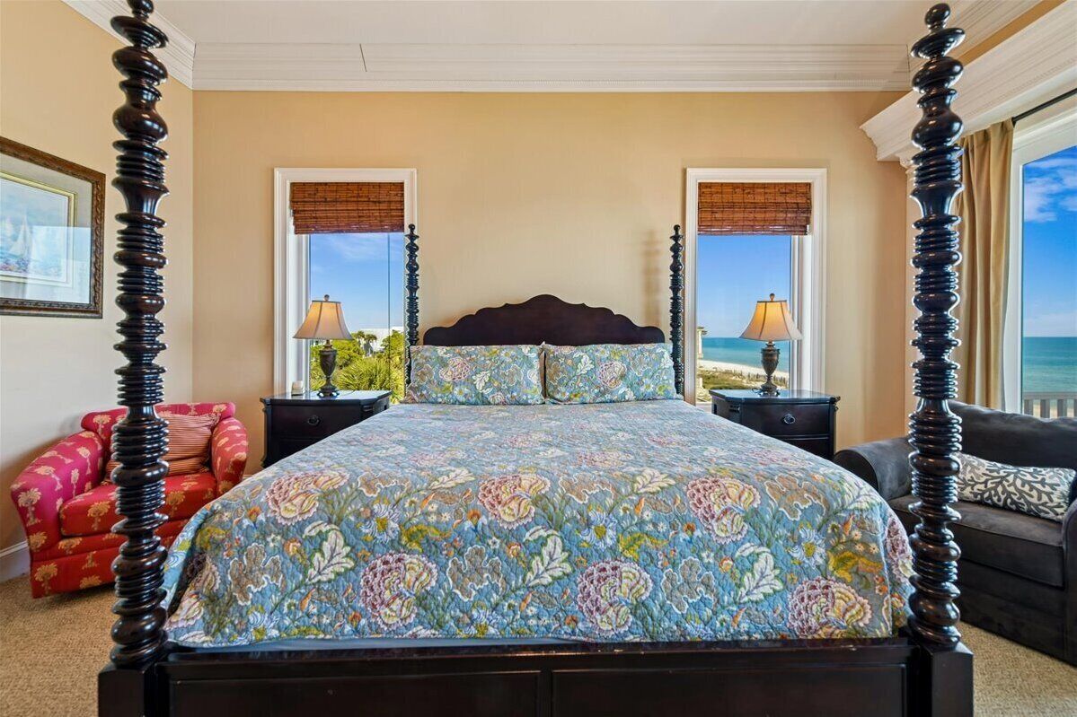 A four-poster bed with a floral bedspread, two bedside tables with lamps, a red armchair, and a view of the ocean through large windows.