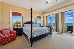 A four-poster bed with a floral bedspread, a red upholstered sofa, and a view of the ocean through large windows.
