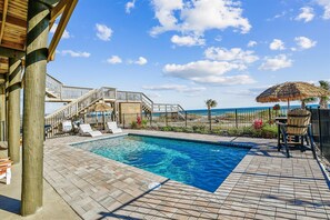 Un espace piscine offrant une vue dégagée sur l’océan, avec une terrasse en bois, des chaises longues et un parasol en chaume.