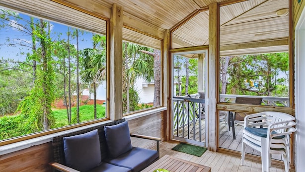 A wooden deck with a sofa and chairs, a dining table, and large windows offering a view of trees and greenery. - Welcome to Sun Treader! (St. George Island)