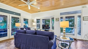 A spacious living room with a ceiling fan, large windows, a blue sofa, and a television. - Welcome to Sun Treader! (St. George Island)