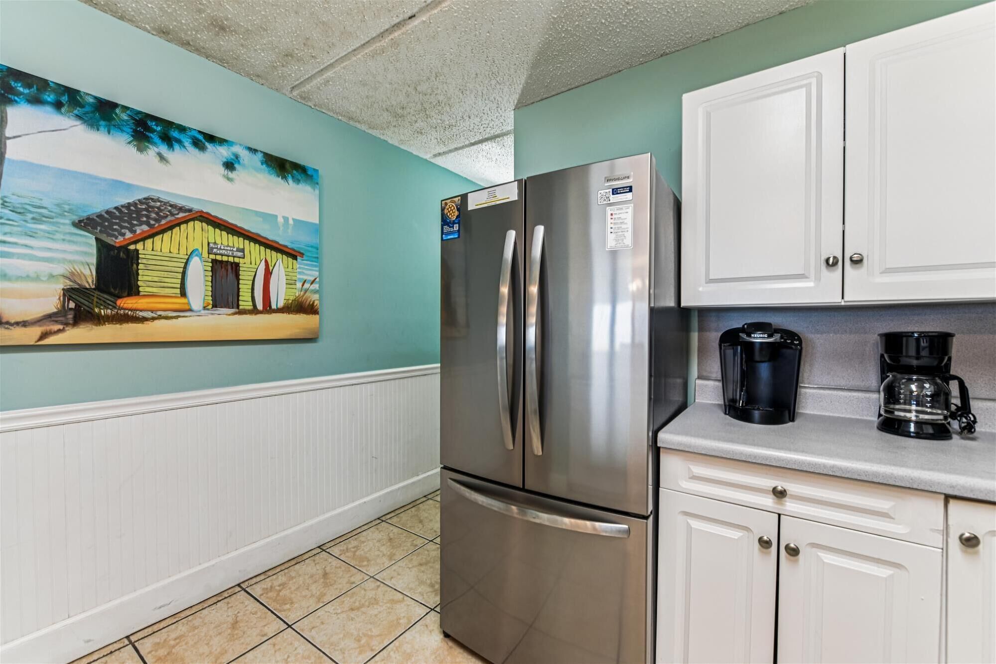 A kitchen with a stainless steel refrigerator, white cabinets, a coffee maker, and a beach-themed painting on the wall.