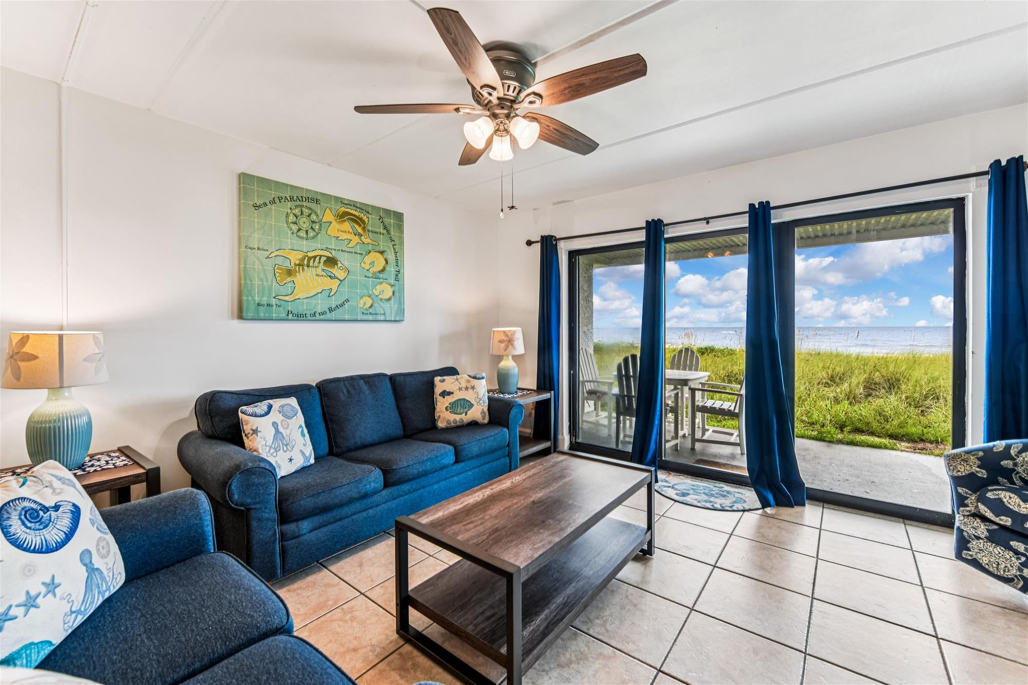 A living room with a blue sofa, a coffee table, and a map on the wall. There is a view of the ocean through large windows.