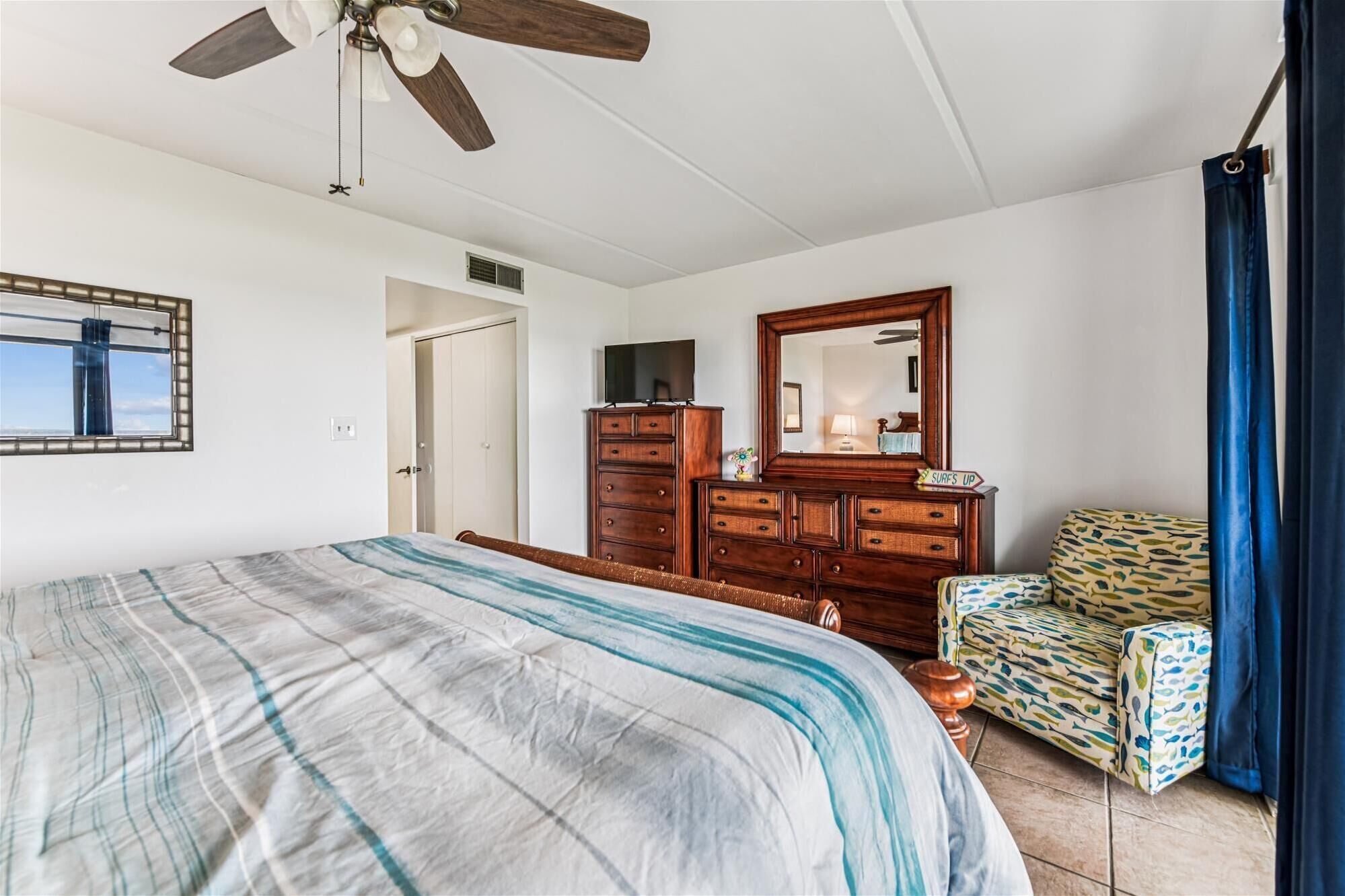 Een slaapkamer met een bed, een dressoir, een stoel, een plafondventilator en een raam met gordijnen.
