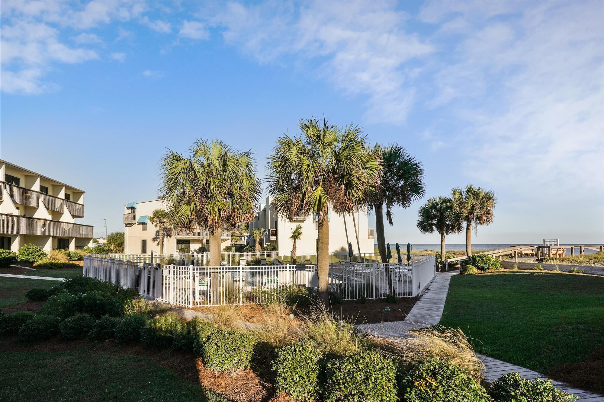 A beachfront hotel with palm trees, a white fence, and a boardwalk leading to the beach.