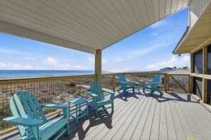 A wooden deck with turquoise Adirondack chairs and a table, overlooking a beach and ocean.
