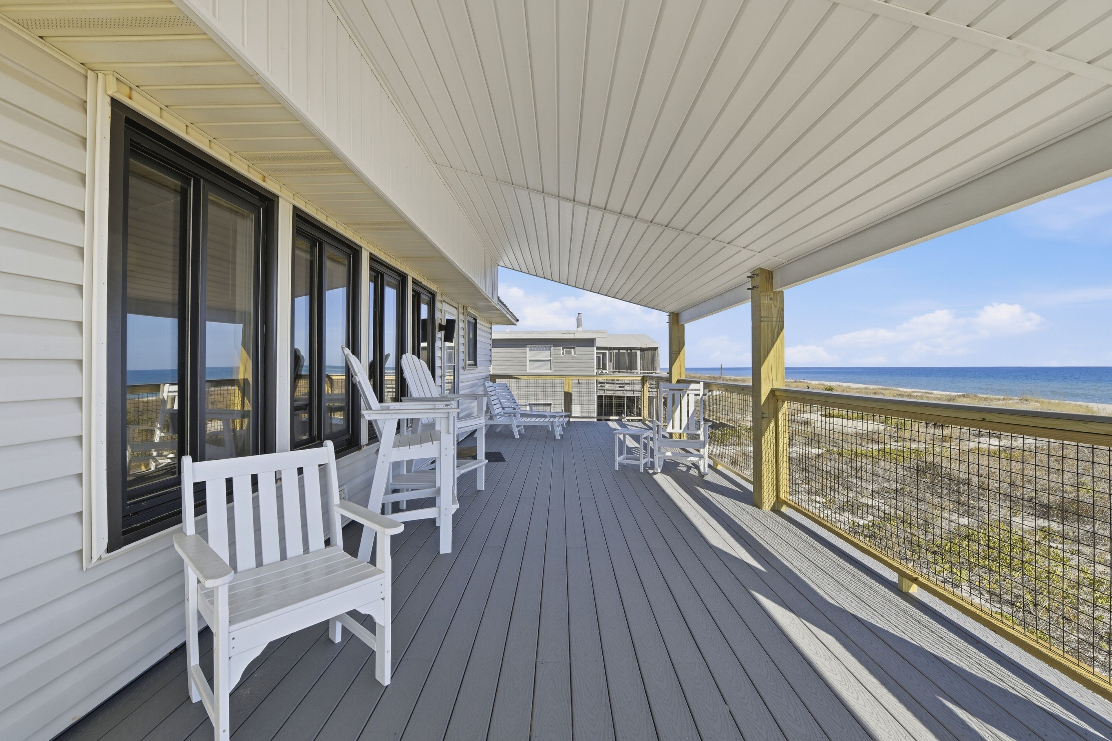 A covered deck with white chairs and a bench, overlooking a beach.