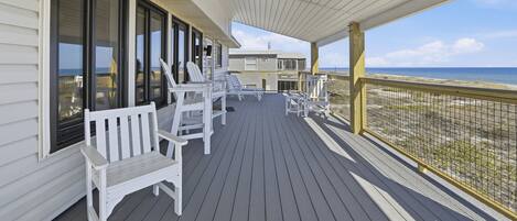 A covered deck with white chairs and a bench, overlooking a beach.