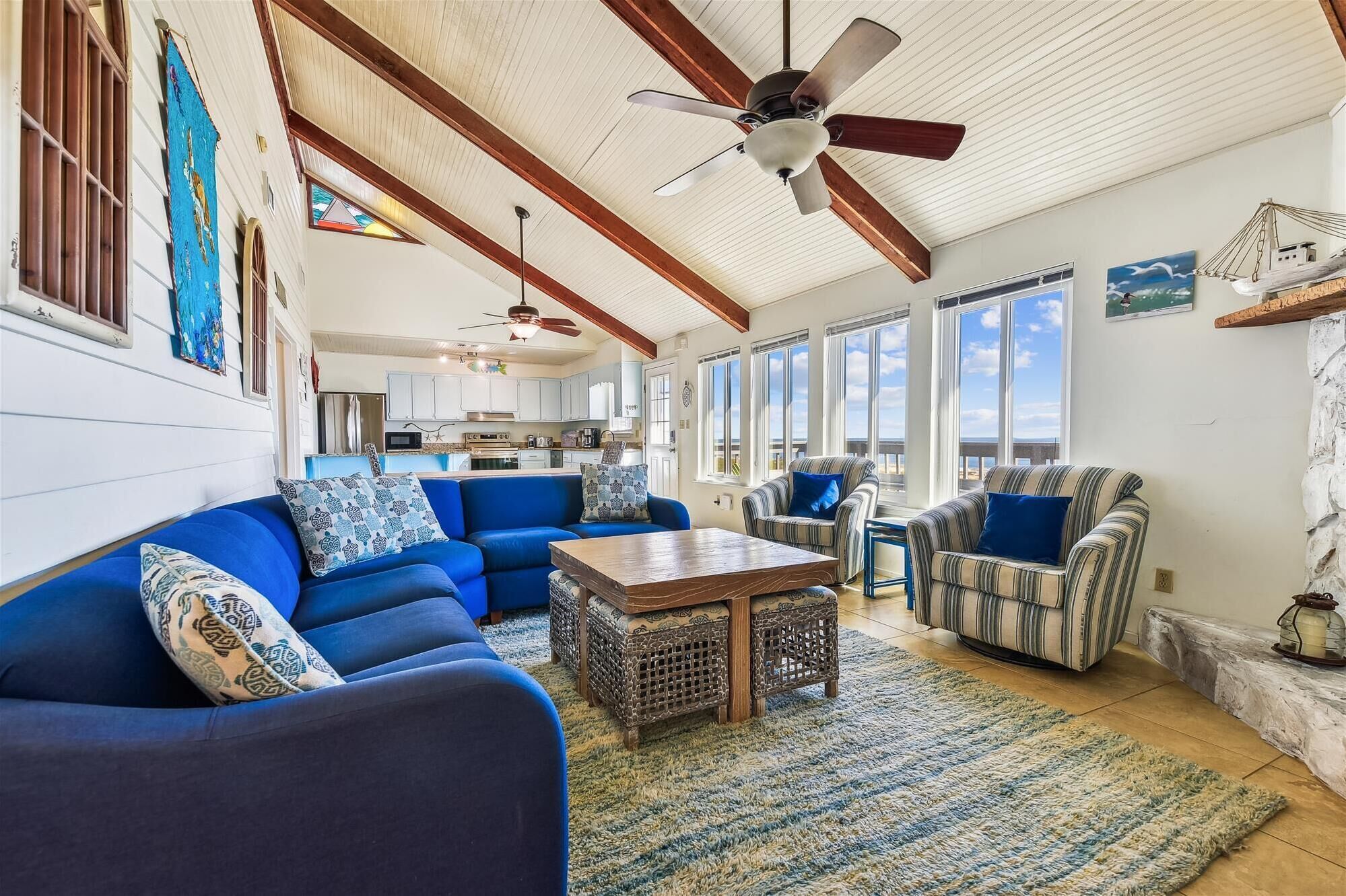A spacious living room with a blue sofa, striped armchairs, and a wooden coffee table. A ceiling fan and large windows provide natural light.