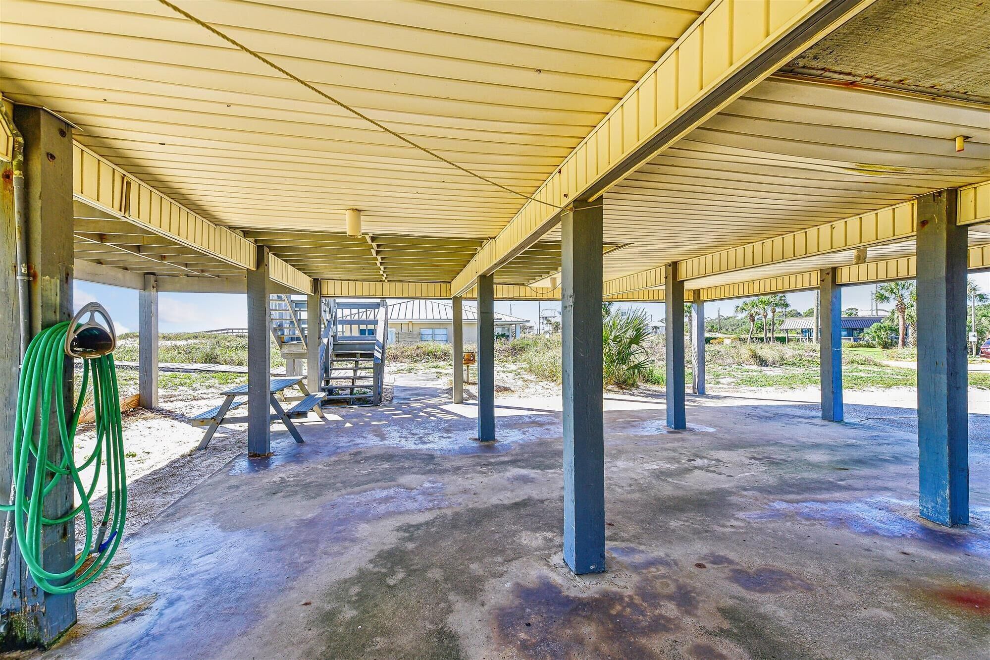 An open-air structure with a concrete floor and metal pillars, featuring a green hose and a staircase leading to an upper level.