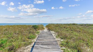 Ein hölzerner Steg führt zum Strand, umgeben von grasbewachsenen Dünen.