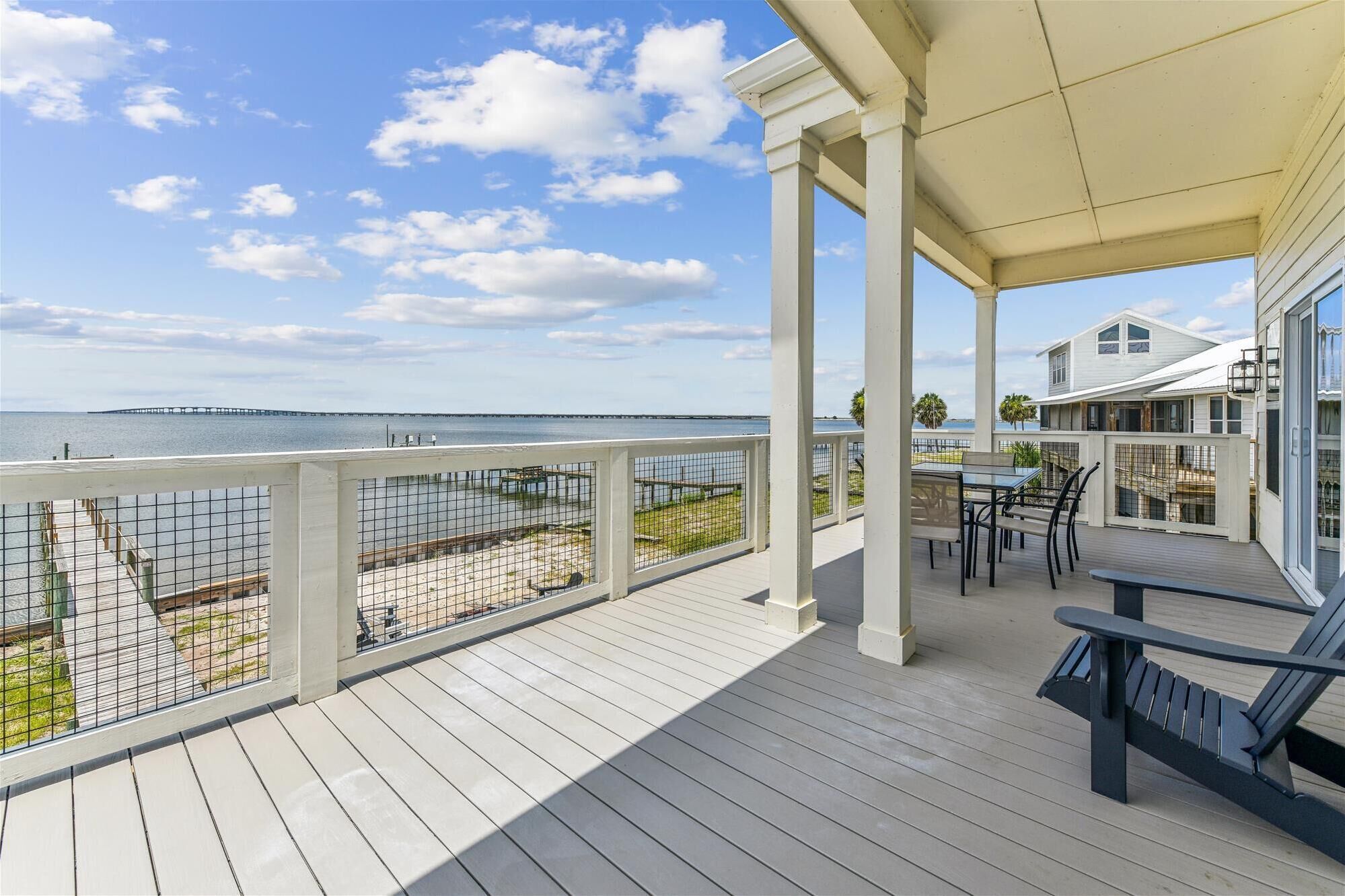 A covered deck with a view of the water, featuring a table and chairs, a railing, and a building in the background.