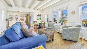 A spacious living room with a blue sofa, a wicker ottoman, and a patterned armchair. A dining area with a table and chairs is visible in the background.