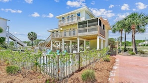 A multi-story beachfront house with a balcony, a staircase leading to the upper levels, and a surrounding garden.