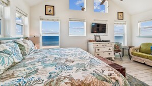 Une chambre dotée d’un grand lit, d’une commode, d’un téléviseur et de plusieurs fenêtres donnant sur l’océan.