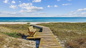 Une promenade en bois mène à un banc situé sur une plage, d’où l’on a une vue dégagée sur l’océan et le ciel.