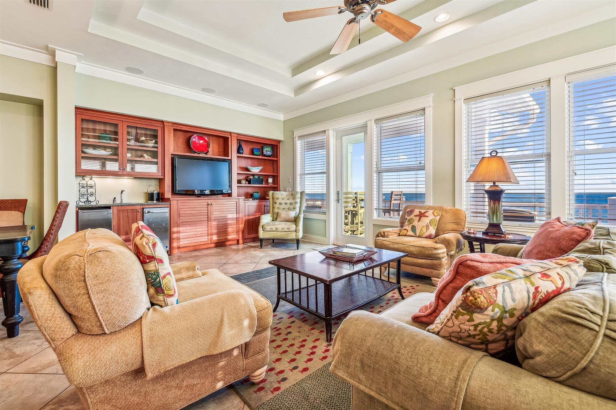A spacious living room with a television, comfortable seating, a coffee table, and large windows with blinds.