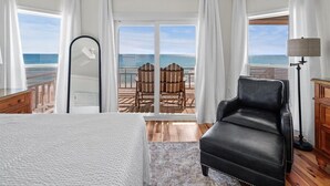 A hotel room with a bed, a black leather chair, a wooden desk, a lamp, and a view of the ocean through large windows. - Welcome to DUNE IT RIGHT! (St. George Island)