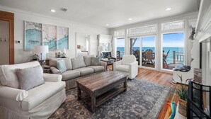 A spacious living room with a large window overlooking the ocean, a white sofa, a wooden coffee table, and a view of the deck. - Welcome to DUNE IT RIGHT! (St. George Island)
