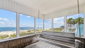 A covered porch with a wooden bench and swing, overlooking a beach with a distant view of a house and palm trees. - Wander from the cool shade of the screened porch to the sun deck with your favorite book! (St. George Island)