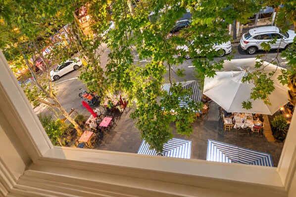 An outdoor dining area with tables and umbrellas, surrounded by trees and a street view.