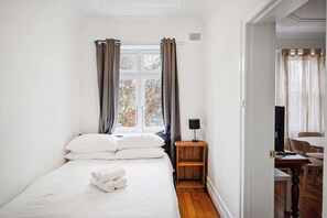 Un dormitorio con cama, ventana con cortinas, mesita de noche con lámpara y vistas al exterior.