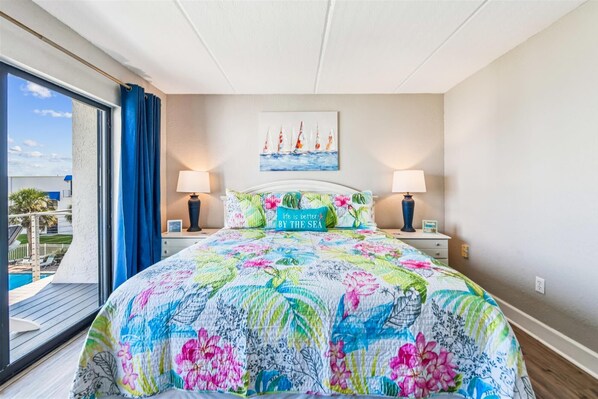 A bedroom with a large bed, two bedside lamps, a painting of sailboats, and a view of a pool and palm trees through a sliding door.