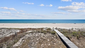 Una playa con un paseo marítimo de madera que conduce a la orilla, arena y un cielo azul despejado.