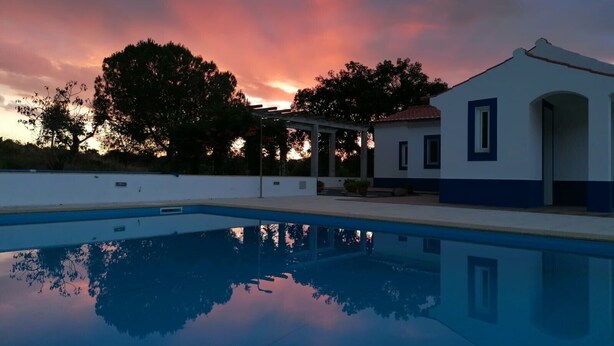 Una piscina con reflejo transparente, un edificio blanco con detalles azules y árboles al fondo.