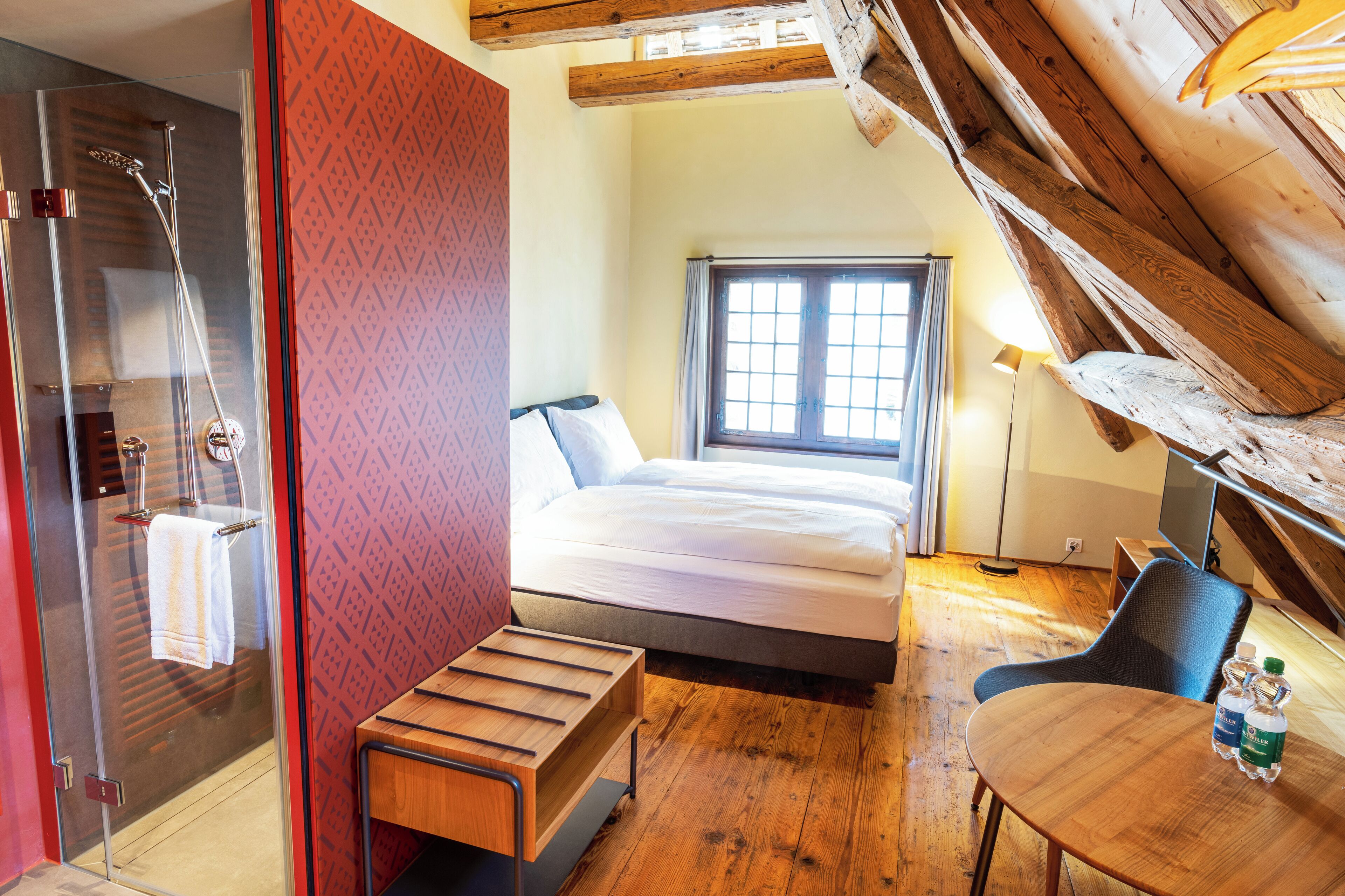 Un dormitorio con cama, un banco de madera, una mesa con botellas de agua, ducha y ventana con cortinas.