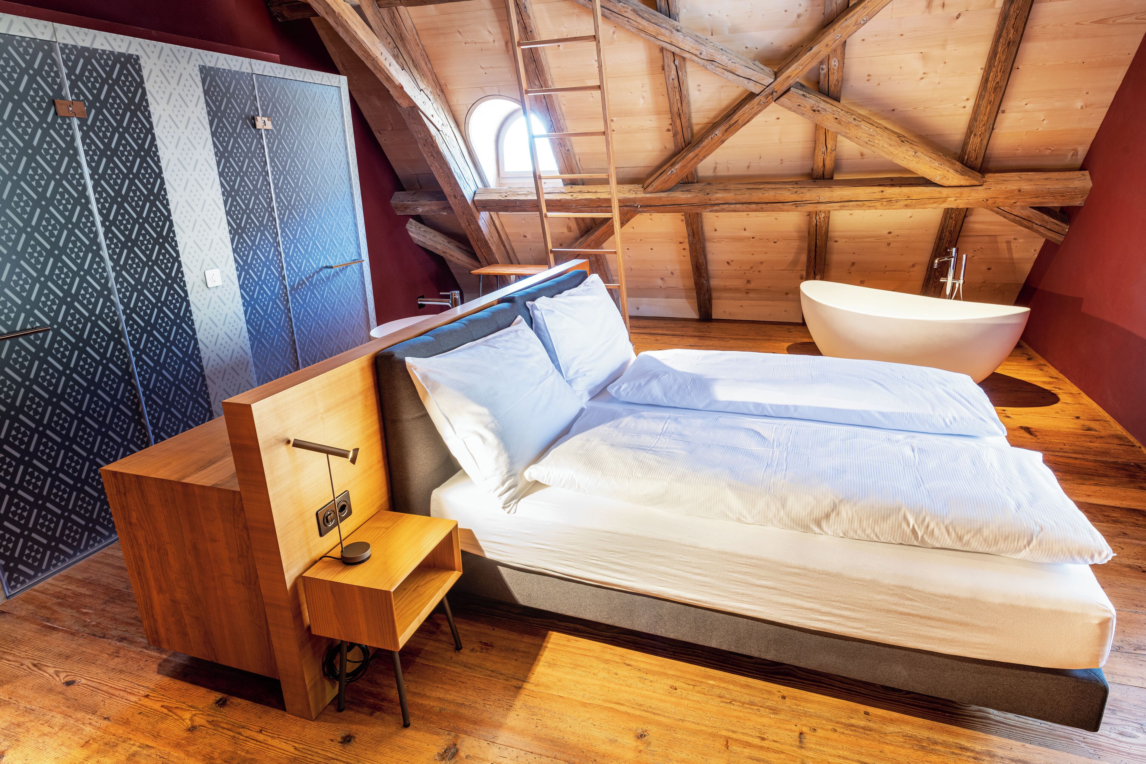 A bedroom with a bed, a wooden nightstand, a built-in shelf, a large window, and a wooden ceiling.