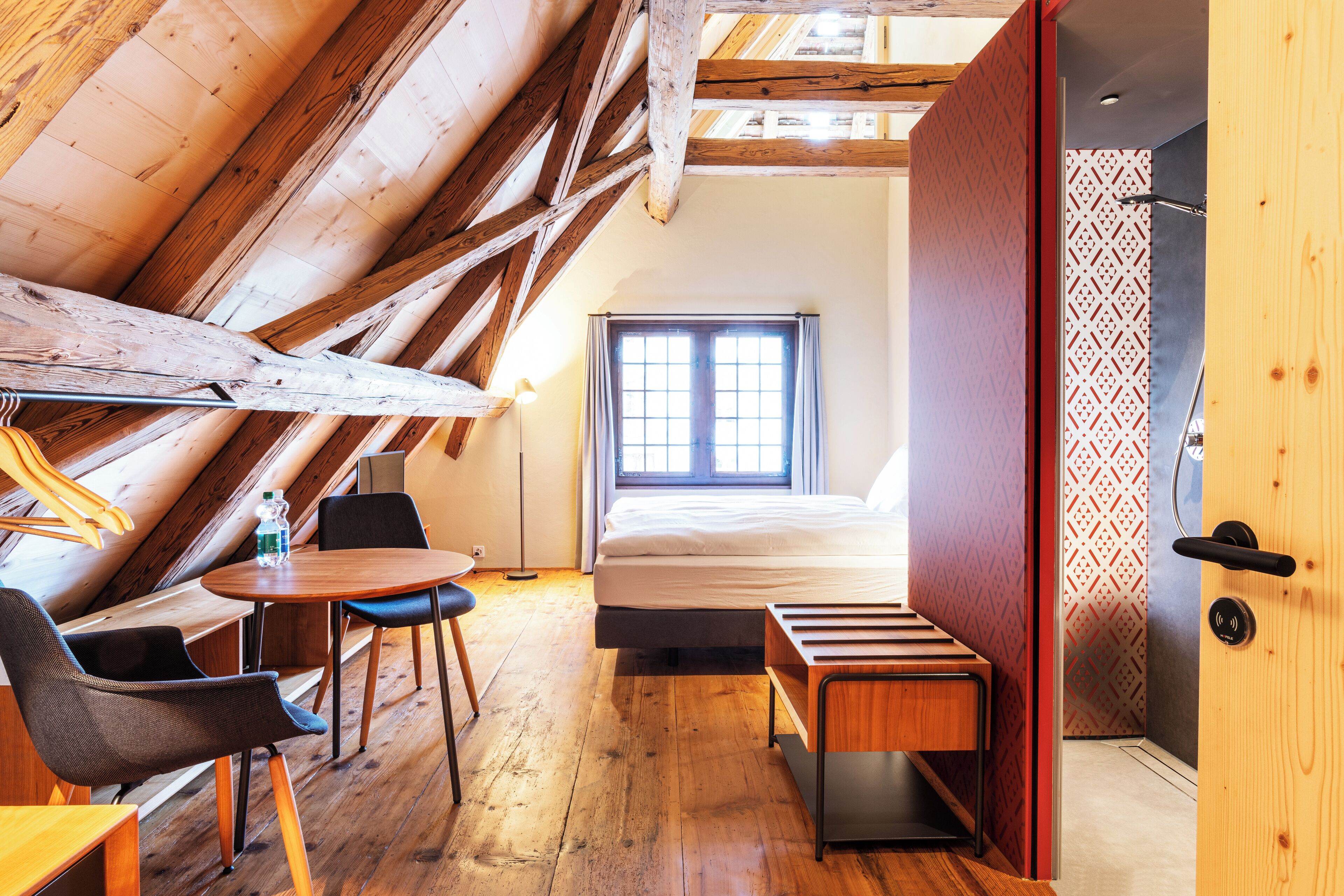 Una habitación con techo inclinado, vigas de madera, cama, ventana, mesa y sillas.