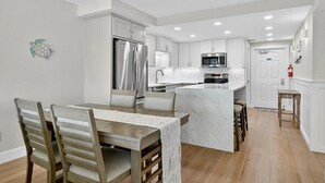 A modern kitchen with white cabinetry, a central island, and a dining area with wooden chairs and a table. - Gulfstream Condos Unit 504 | Pool, Hot Tub & BBQ Areas | Private Balcony (Corpus Christi)
