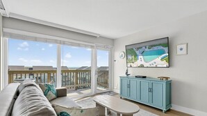 A spacious living room with a large sliding glass door leading to a balcony, a teal cabinet, and a flat-screen TV mounted on the wall. - Gulfstream Condos Unit 504 | Pool, Hot Tub & BBQ Areas | Private Balcony (Corpus Christi)