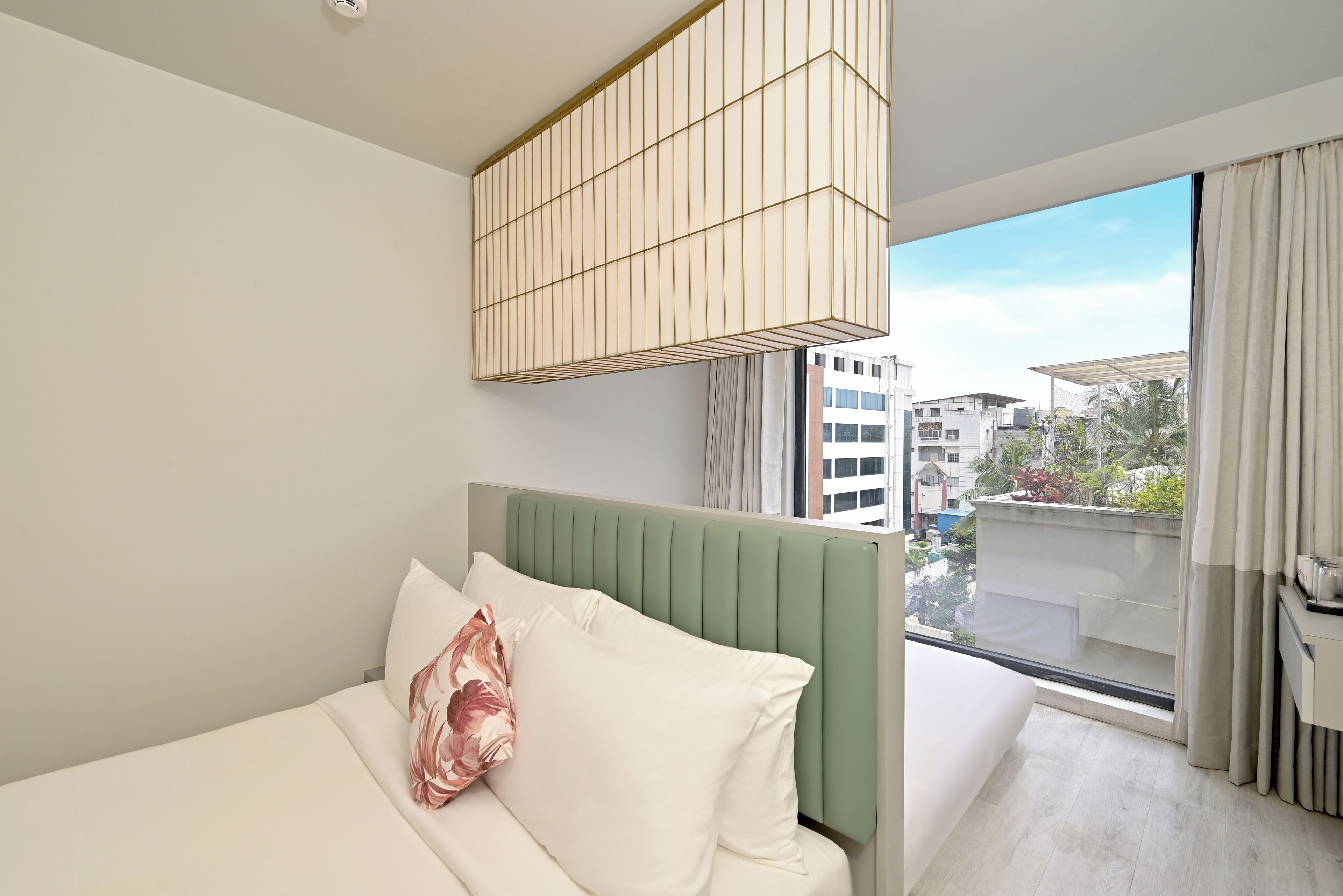 A modern hotel room with a bed, a green headboard, a hanging light fixture, and a view of buildings through a large window.