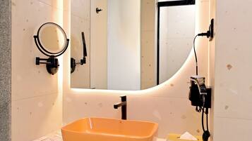 Une salle de bain avec un mur recouvert de carreaux blancs, un grand miroir, un miroir rond, un lavabo orange, un porte-serviettes et une boîte à mouchoirs.