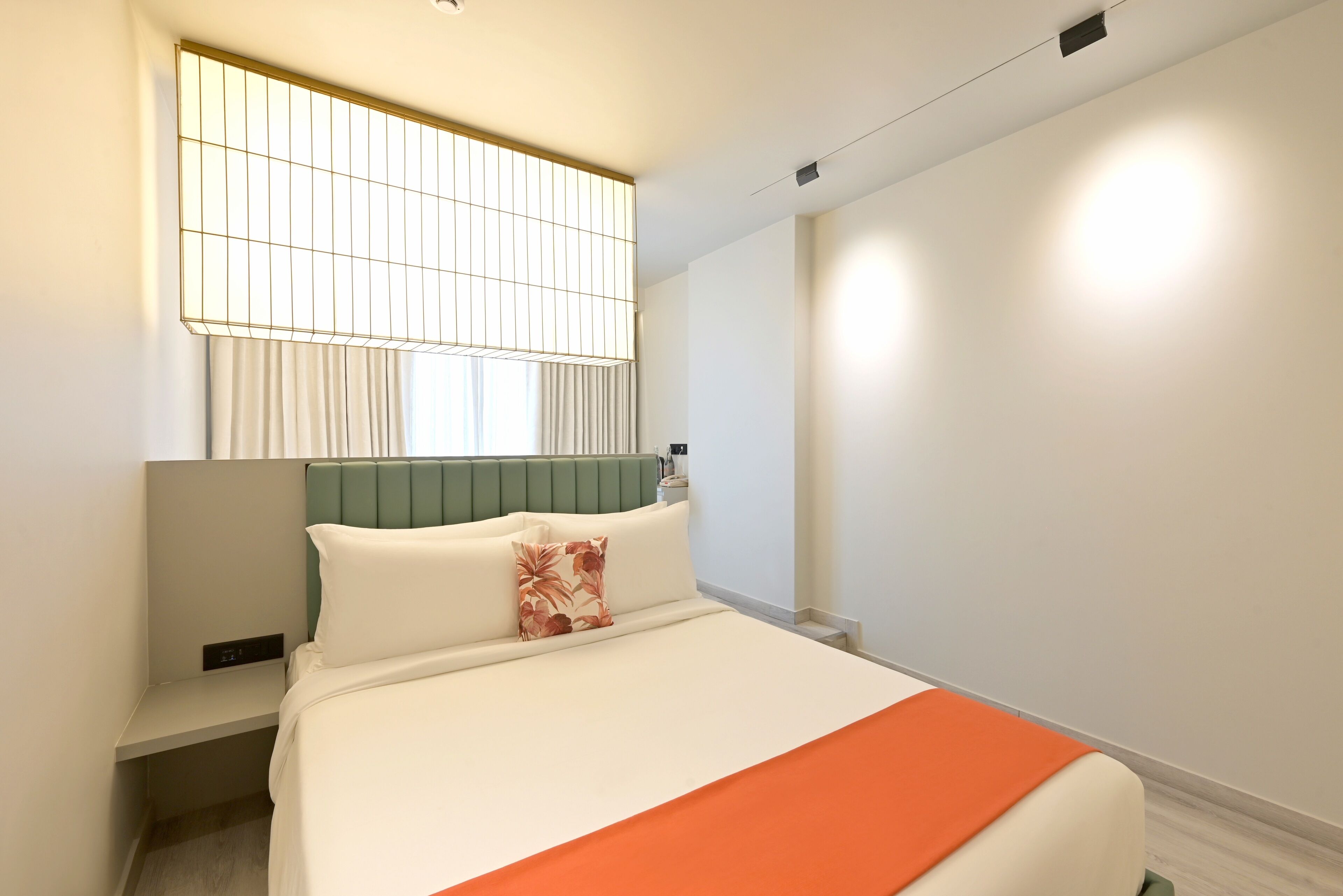 A modern hotel room with a bed, a nightstand, a window with blinds, and a wall-mounted light fixture.