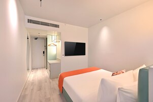A modern hotel room with a bed, a TV mounted on the wall, and a hallway leading to a bathroom.
