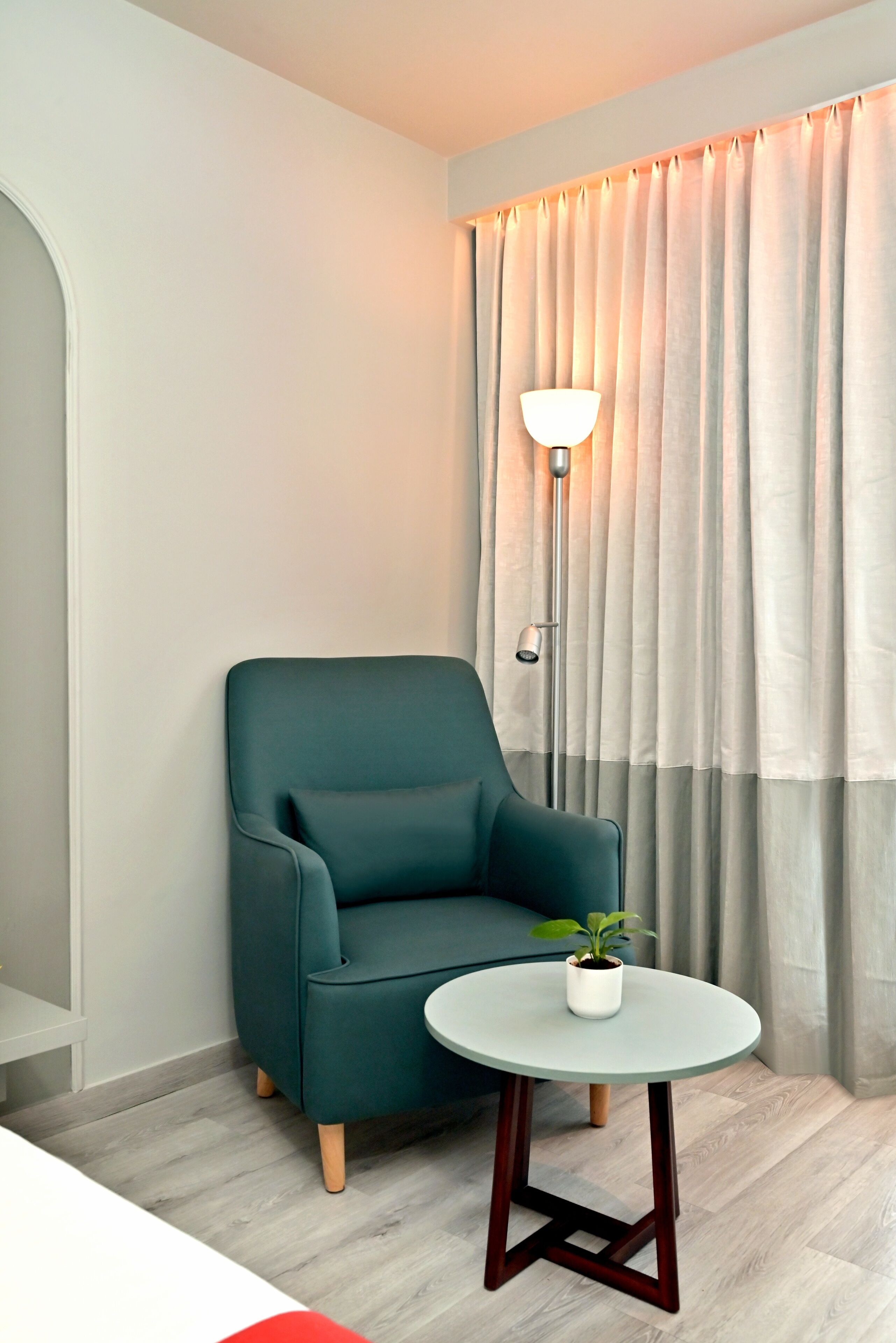 A modern room with a teal armchair, a small round table with a potted plant, a floor lamp, and a large window with curtains.
