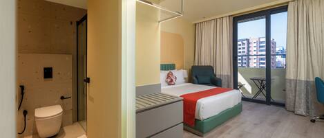Habitación de hotel con cama, una silla, una mesita, balcón con vista a edificios y baño con inodoro y ducha.