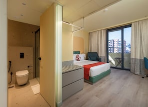Habitación de hotel con cama, una silla, una mesita, balcón con vista a edificios y baño con inodoro y ducha.