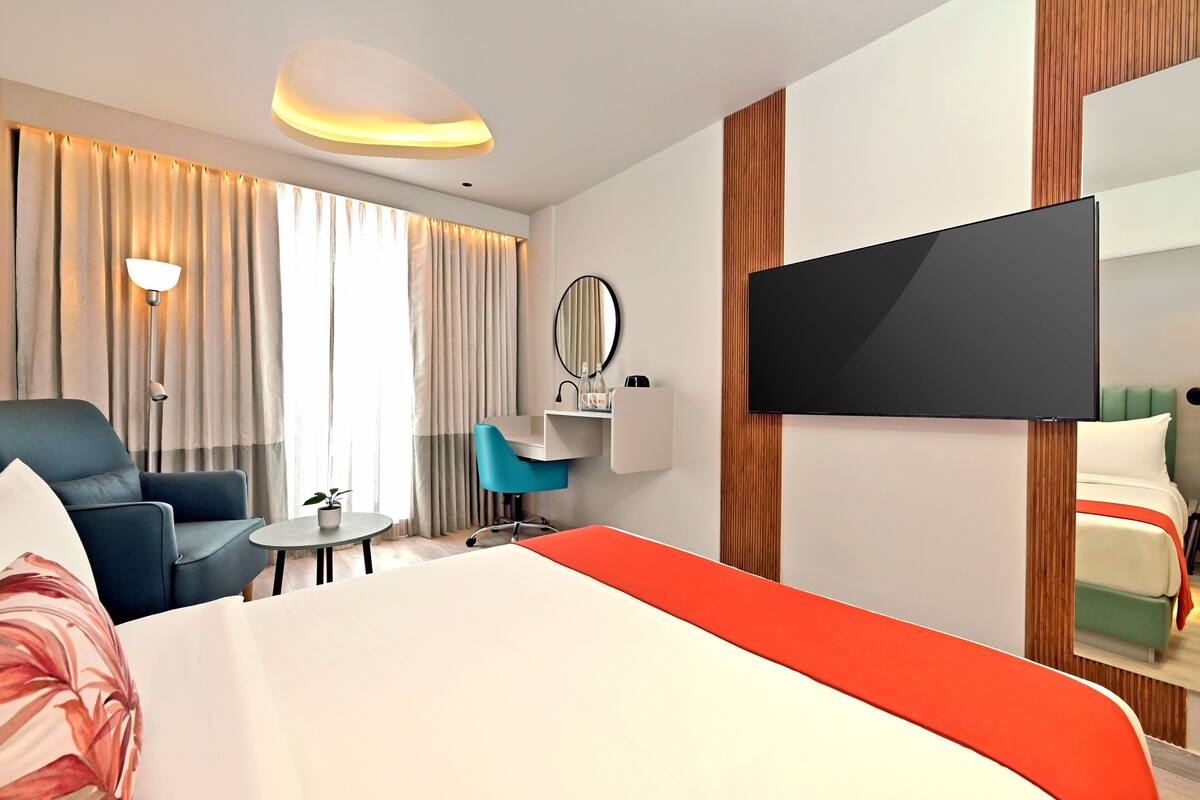 Habitación de hotel con una cama grande, un escritorio con silla, una mesita con planta, un televisor montado en la pared y una ventana con cortinas.