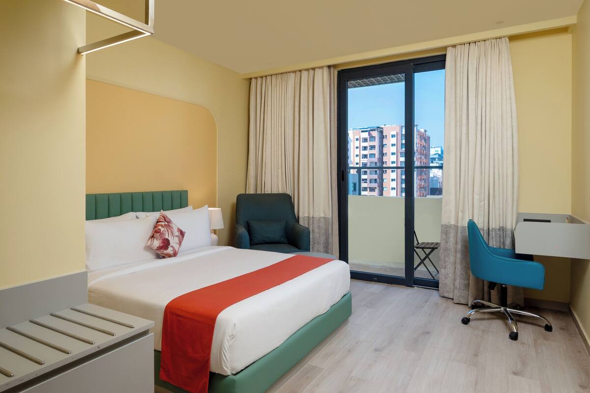 Habitación de hotel con cama, escritorio, silla y vista a edificios a través del ventanal.